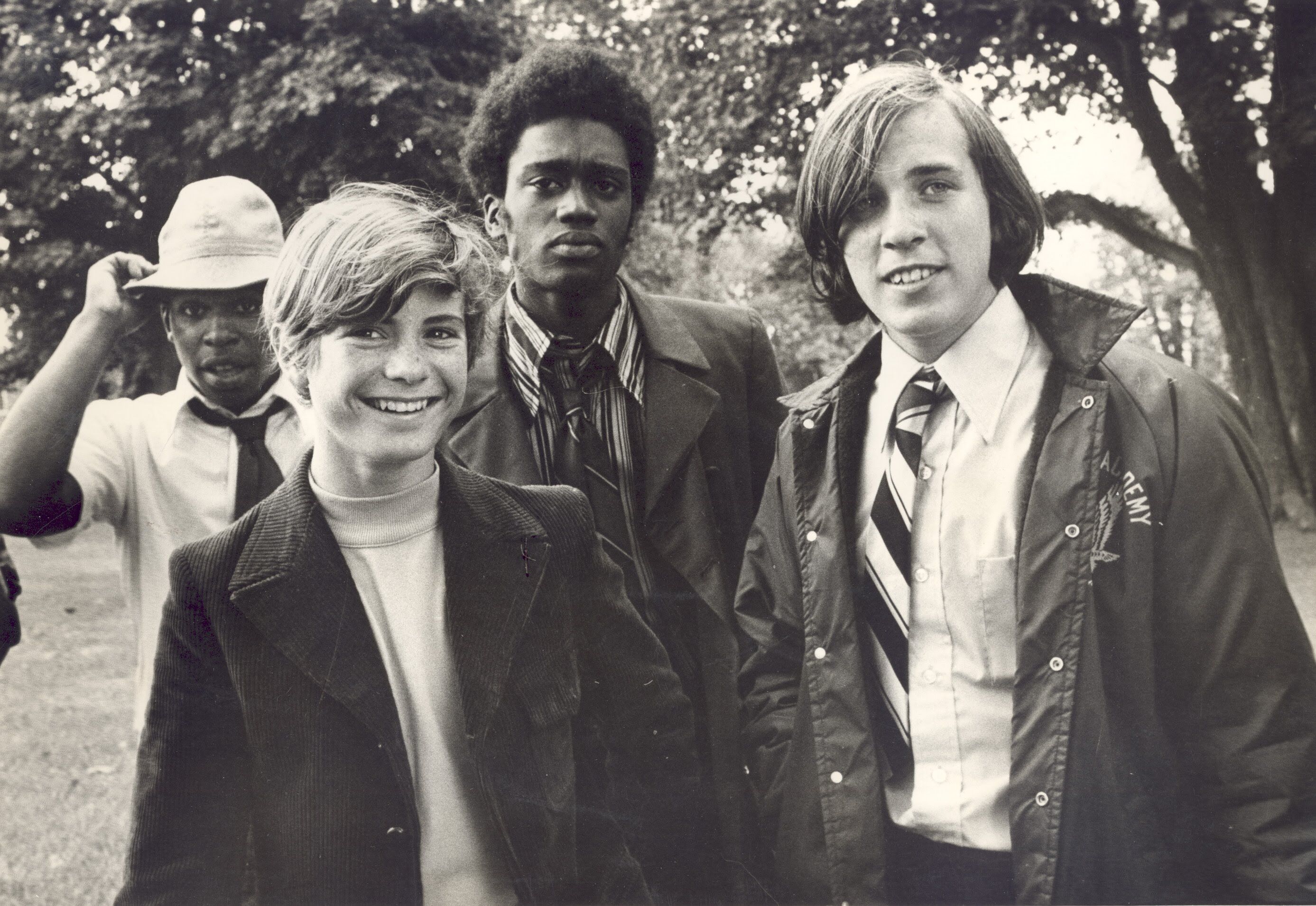 The image shows a group of four young men posing outdoors. They appear to be from the 1970s, given their clothing and hairstyles. The men are of different ethnicities and seem to be friends. They are smiling and looking at the camera. The background is blurred, suggesting a park or outdoor setting.